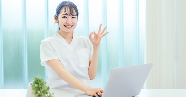 白衣姿の女性、パソコンの前でOKサインを出している。