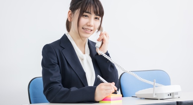 リクルートスーツ姿で、座りながらメモを片手に電話をかける女性。