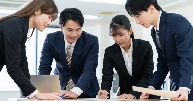 スーツ姿の男女４人。デスクで発表資料を共同作業中。