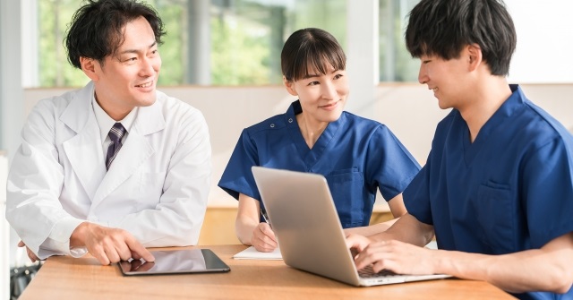 白衣姿の３人の男女。机の上でパソコンで作業中。