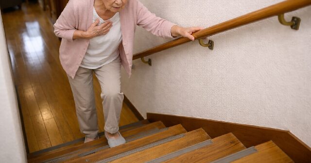 自宅の階段を昇る高齢者の女性。手摺りを持って不安そうな表情。
