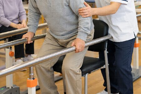 リハビリ室で訓練中の高齢者。理学療法士に支えられ平行棒をつかんで立ち上がり訓練中。