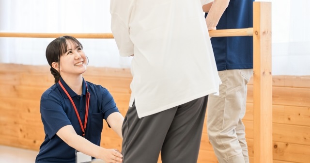 リハビリ室で手すりをつかって歩く高齢者。介助する医療従事者